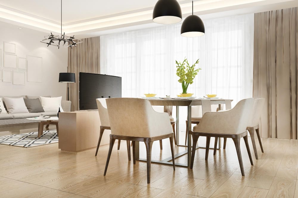 Open-concept living and dining area with six beige upholstered chairs around a rectangular dining table, large windows with sheer curtains, and a sofa in the background.