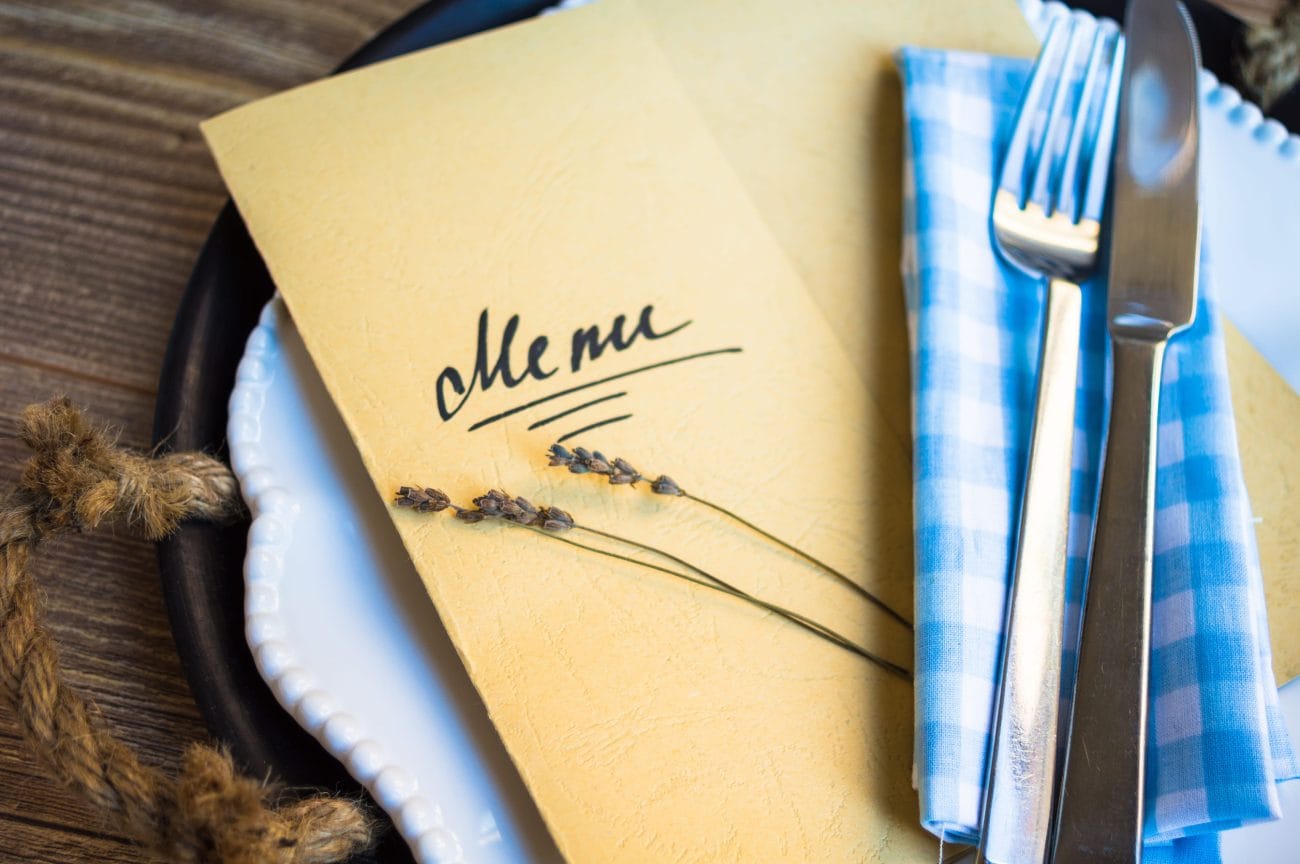 Menu card on a rustic wooden table with lavender sprigs, a white plate, and a blue checkered napkin with knife and fork nearby.