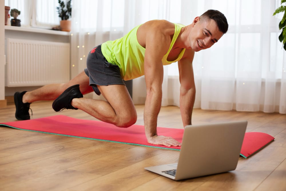 Man in a neon tank top on a pink yoga mat performs a mountain climber exercise while following an online workout on a laptop placed on the floor at home.