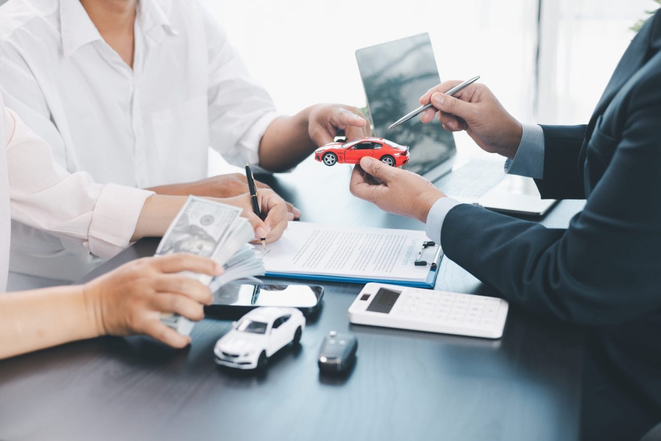 Two professionals negotiate auto financing at a desk, exchanging a red toy car model with money and documents nearby on the table.