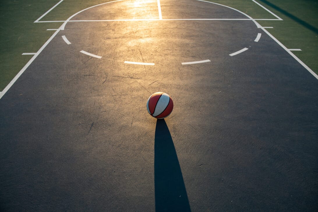 775540 Mtzusa - Hunter Magazine Basketball sits at the center of an outdoor court with a long shadow extending toward the camera as sunlight glints off the surface.
