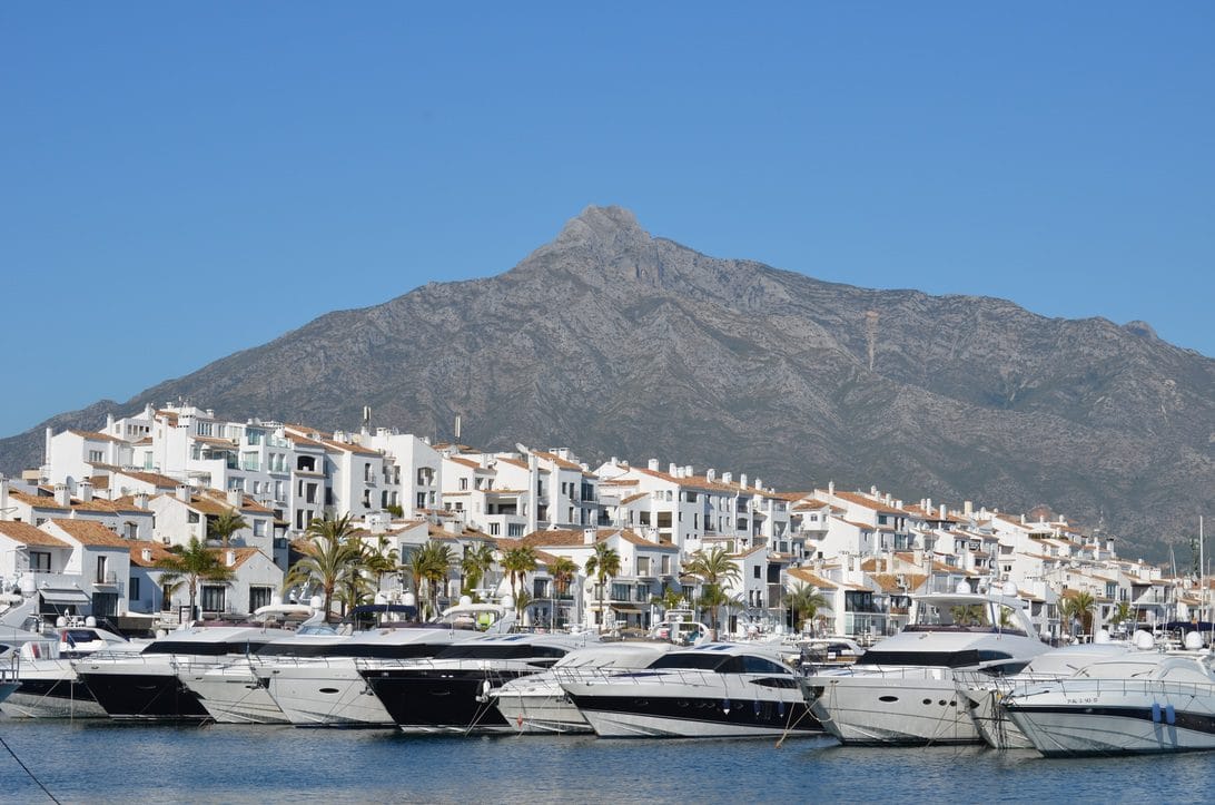 Row of white Mediterranean houses with red-tiled roofs along a marina, moored yachts, and a mountain backdrop (clear blue sky).