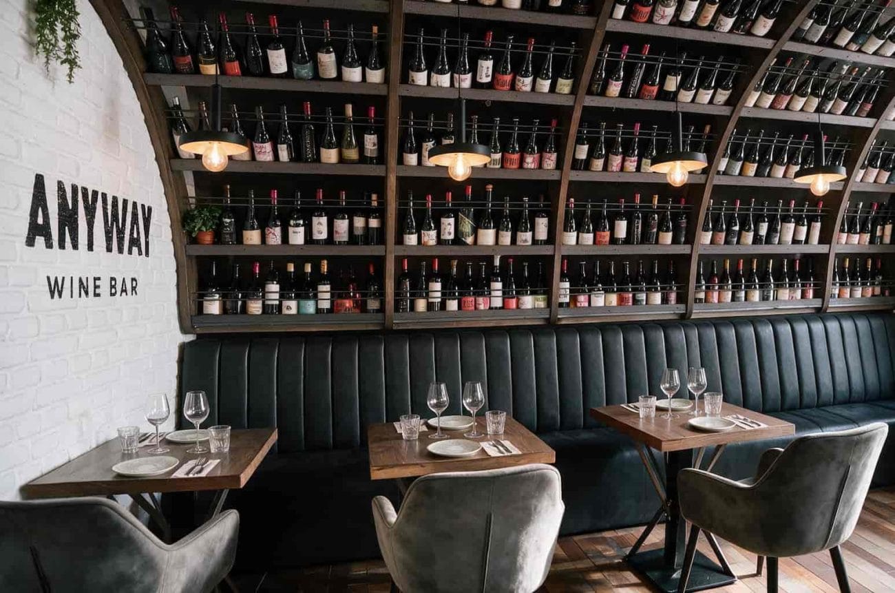 Cozy wine bar with a long green upholstered banquette, wooden tables, and shelves full of wine bottles along a white brick wall labeled Anyway Wine Bar.