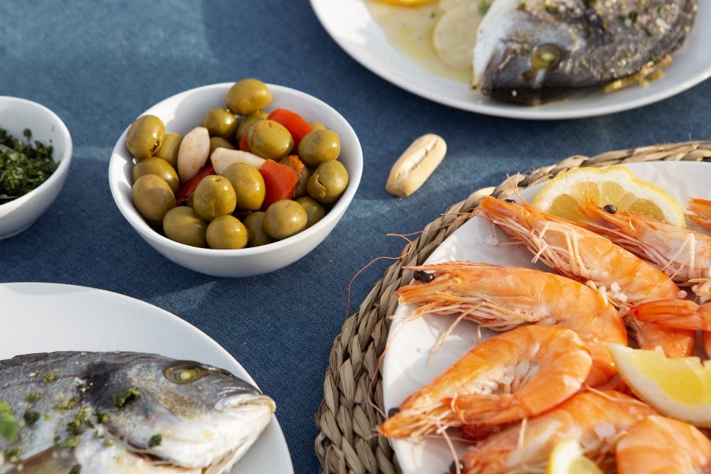 778927 - Restaurante Las Palmas - Hunter Magazine Plate of cooked shrimp with lemon wedges on a woven placemat, beside bowls of olives and whole fish on a blue tablecloth.