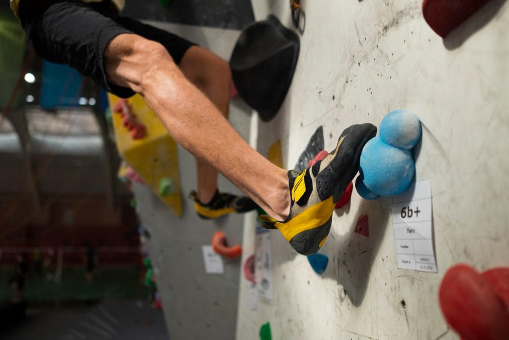 779249 - ECO CLIMB - Hunter Magazine Climber's foot in a yellow-and-black climbing shoe on a blue hold at an indoor rock climbing wall; route label '6b+' visible.
