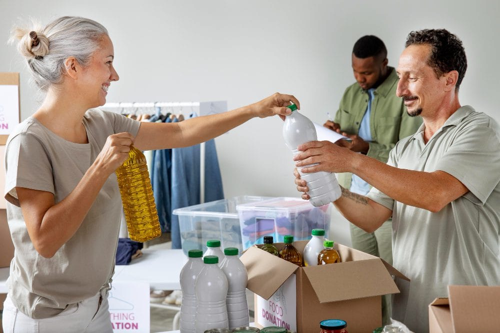 780380 - Cáritas Zaragoza - Hunter Magazine Woman hands a bottle of oil to a man at a donation center; they smile as they exchange items with boxes of supplies nearby.