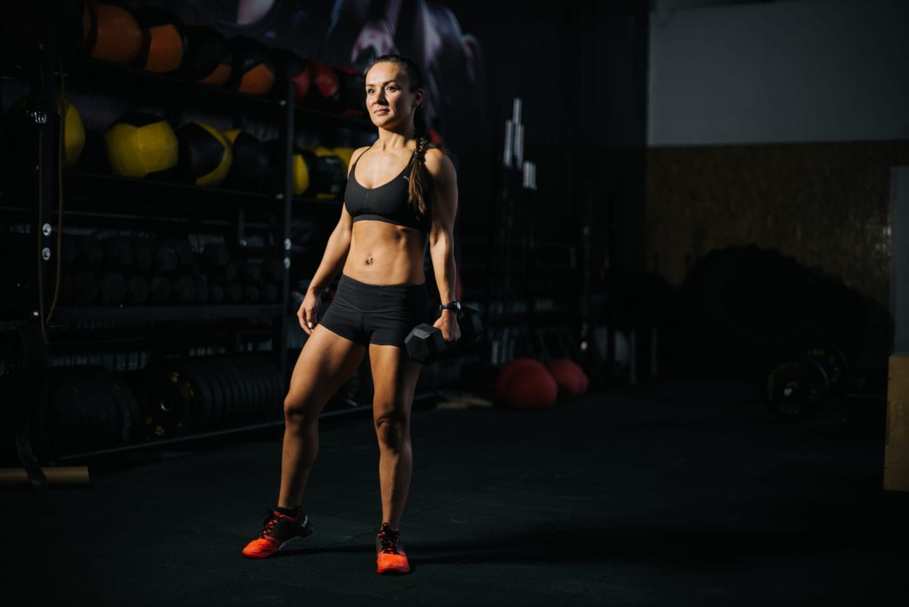 Athlete woman in a black sports bra and shorts stands in a dim gym, holding a dumbbell in one hand with equipment in the background.