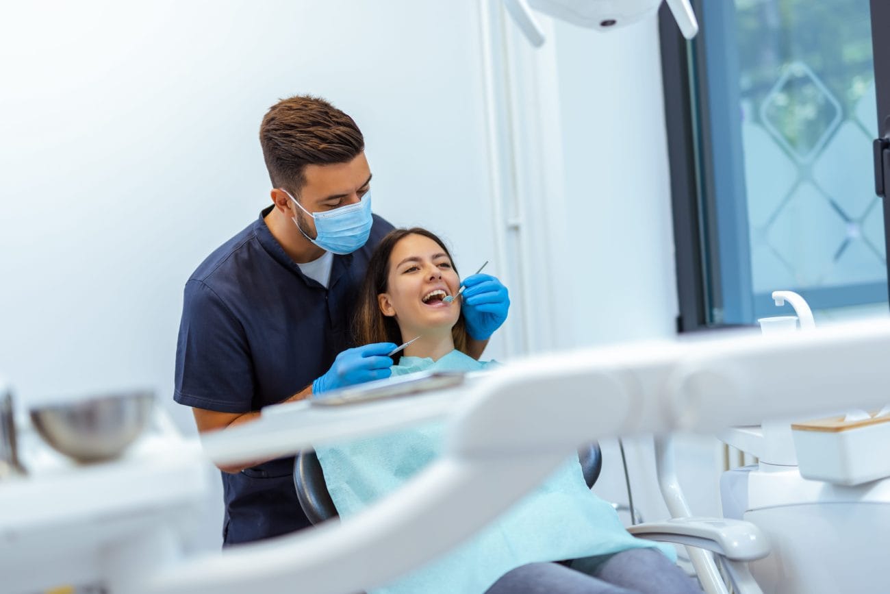 Dental professional wearing a mask and gloves examines a patient's teeth in a bright dental clinic.